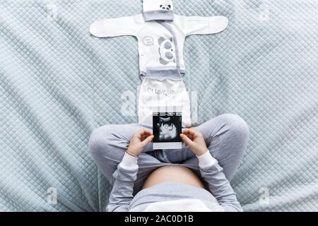 Femme enceinte attend de toucher son ventre tout en regardant l'échographie, assis sur un lit confortable avec des couvertures, oreillers, vêtements d'enfants. Banque D'Images