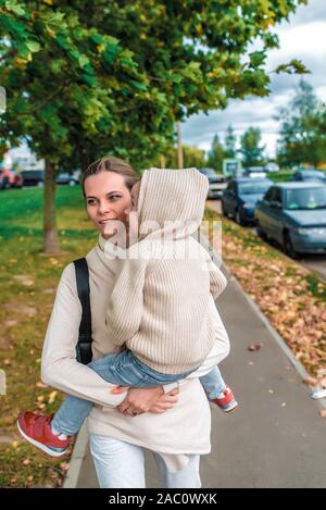 Femme mère marche le long de la rue, est titulaire d'petit garçon fils ses bras, ville d'automne, à pied le long de rue, arbres en arrière-plan, des vêtements chauds, pull beige Banque D'Images