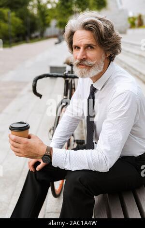 Photo de serious mature businessman dans des vêtements à emporter boire du café tout en étant assis sur un banc de travail avec location en plein air Banque D'Images