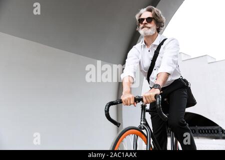 Photo de confiance mature businessman en lunettes de soleil et chemise blanche riding bicycle on city street Banque D'Images