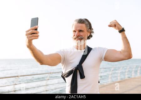Image of old man in sportswear montrant ses biceps tout en prenant des photos sur selfies at promenade de matin Banque D'Images