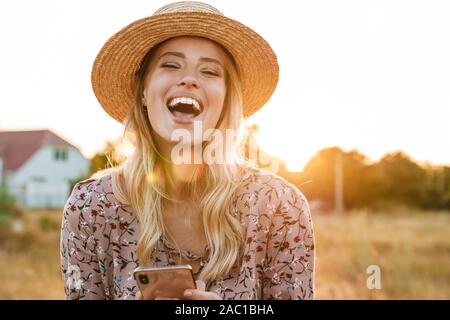 Photo de young blonde woman wearing straw hat holding cellulaire et un rire tout en marchant dans la campagne environnante au cours de journée ensoleillée Banque D'Images