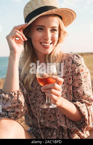 Photo de cheerful Pretty woman in straw hat holding wine glass et souriant tout en restant assis près de station au cours de journée ensoleillée Banque D'Images