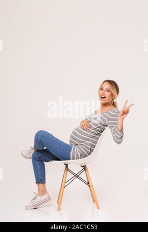 Jolie jeune femme enceinte assise sur une chaise isolée sur fond blanc, montrant le geste de paix Banque D'Images