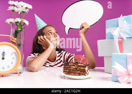 Jolie jeune fille assise au bureau avec un gâteau d'anniversaire sur isolé sur fond rose, la célébration, la holding empty speech bubble Banque D'Images