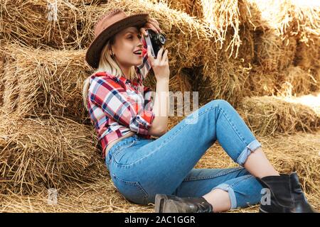 Smiling young blonde cowgirl assis sur une botte de foin dans la grange, prendre des photos avec l'appareil photo Banque D'Images