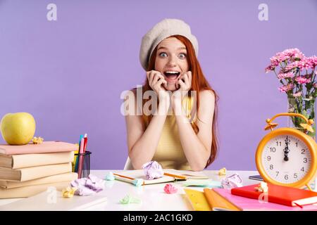 Photo de jeune fille étudiante excitée assis à un bureau avec les livres d'exercice tout en faisant ses devoirs sur fond violet isolés Banque D'Images