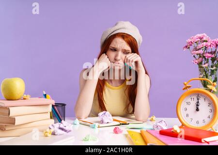 Photo de jeune fille étudiante de pleurer assis à un bureau avec les livres d'exercice tout en faisant ses devoirs sur fond violet isolés Banque D'Images