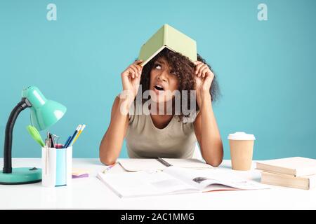 Belle jeune femme africaine bouleversé habillé négligemment assis au bureau isolé sur fond bleu, holding book sur sa tête Banque D'Images