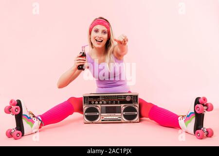 Portrait d'une jeune fille blonde portant des vêtements de sport rétro et de patins assis plus isolé sur fond rose avec boombox, holding bo Banque D'Images