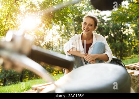 Belle Jeune Fille habillée négligemment assis sur un banc à côté de son scooter au city park, laughing Banque D'Images