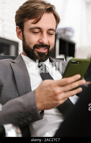 Photo de concentrés d'affaires en costume officiel de la saisie sur cellphone while sitting in bureau open space Banque D'Images