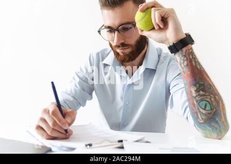 Image of young man wearing eyeglasses holding tennis ballon en working in office Banque D'Images