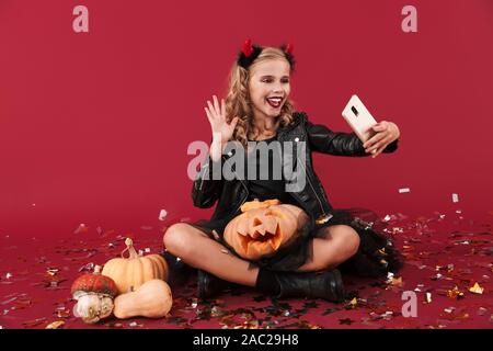 Photo de smiling happy little girl positive devil in carnival costume halloween isolé sur fond mur rouge holding pumpkin parle par mobile Banque D'Images