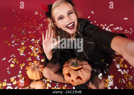 Photo de petite fille optimiste en diable carnival costume halloween isolé sur fond mur rouge holding pumpkin prendre forme par caméra selfies t Banque D'Images