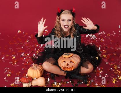 Photo de petite fille heureuse joyeuse dans devil costume halloween carnaval sur fond Mur isolé rouge holding pumpkin. Banque D'Images