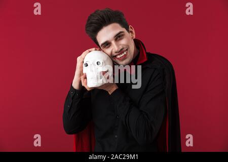 Image homme crocs de vampire avec en noir halloween costume holding crâne humain isolated over red wall Banque D'Images