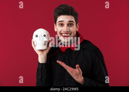 Image de l'homme vampire excité avec crochets en noir halloween costume holding crâne humain isolated over red wall Banque D'Images