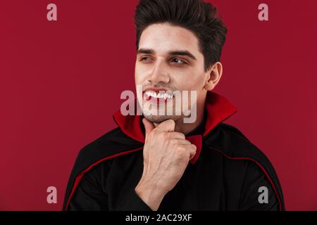 Image de l'homme vampire joyeux avec du sang et des crocs en noir costume halloween isolated over red wall Banque D'Images