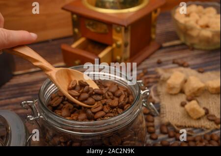 La main de la jeune fille prend une cuillère en bois de grains de café à partir d'un bocal en verre, un moulin à café, morceaux de sucre brun sur un fond de bois. Close up Banque D'Images
