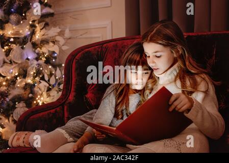 Heureux les jeunes sœurs de la lecture d'un livre d'histoires réunies par une cheminée dans un confortable salon sombre la veille de Noël. La célébration de Noël à la maison. Banque D'Images