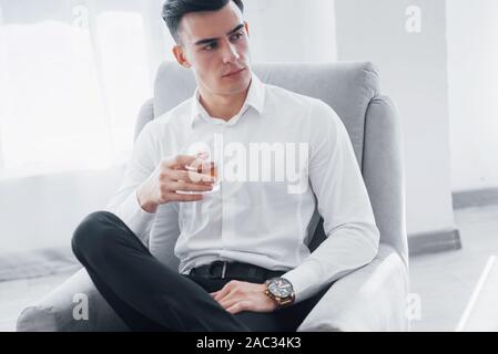 À la recherche sur le côté. Vue en gros plan. Les jeunes gars en élégant costume blanc assis sur la chaise et détient avec l'alcool en verre Banque D'Images