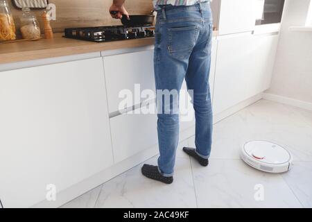 Robot-aspirateur nettoie blanc marbre dans la cuisine tandis que l'homme est la cuisson. Banque D'Images