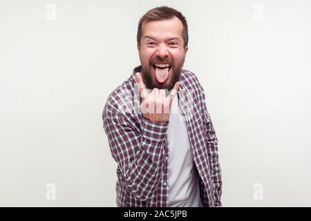 Portrait d'homme barbu optimiste fou en chemise à carreaux montrant rock and roll sign hand gesture et sticking out tongue, sentiment d'être cool en bascule. Banque D'Images