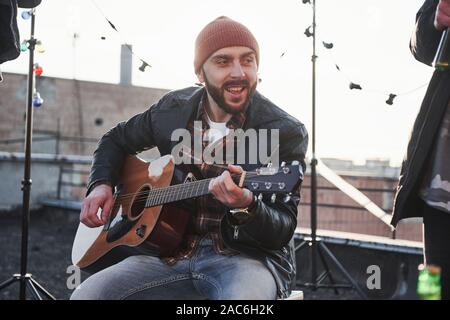 Jeune homme barbu. Guitare acoustique avec Guy chante. Les amis s'amuser at rooftop party avec des ampoules colorées Banque D'Images
