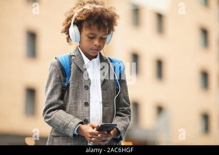 Écolier africain dans les écouteurs d'écouter de la musique sur son téléphone portable tout en se tenant à l'extérieur Banque D'Images