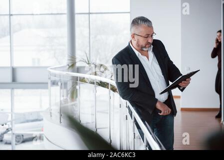 Est subordonné et va avoir une conversation par téléphone mobile derrière. Photo de senior businessman dans la chambre spacieuse avec des plantes. La tenue et la lecture Banque D'Images