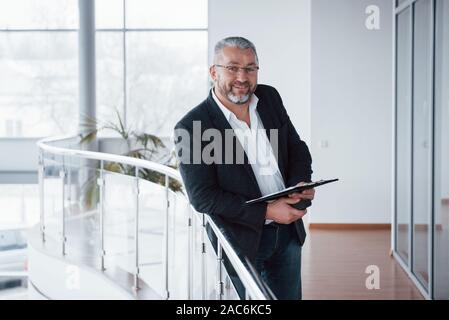 Manager avec succès. Vous pouvez le voir par son sourire sincère. Photo de senior businessman dans la chambre spacieuse avec des plantes derrière. La tenue et la lecture Banque D'Images