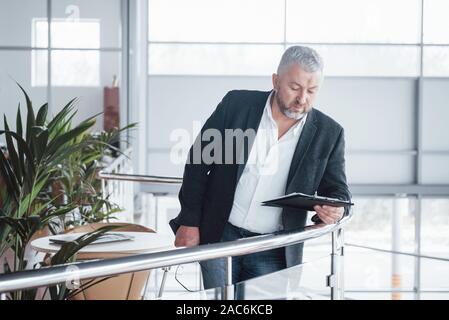 Photo de senior businessman dans la chambre spacieuse avec des plantes et la table en elle. La tenue et la lecture de documents Banque D'Images