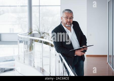 Bon portrait. Photo de senior businessman dans la chambre spacieuse avec des plantes derrière. La tenue et la lecture de documents Banque D'Images