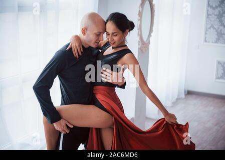 Jolie jeune femme en rouge et noir danse avec chauve dans la chambre blanche Banque D'Images