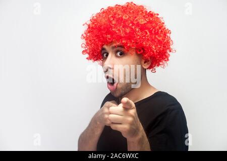 Hey you ! Portrait de l'homme drôle de surprise à poils bouclés et avec perruque rouge pointant à l'appareil photo et à la recherche avec choqué étonné face hilarante expression. Banque D'Images