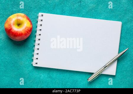 Carnet spirale blanc avec une pomme et un stylo contre le papier texturé artisanal turquoise Banque D'Images