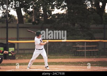 Un dix-huit ans homme balançant le bat à la plaque dans un livre blanc et rouge dans un uniforme de baseball baseball jeu d'été Banque D'Images