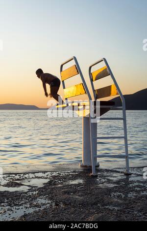 Garçon saute sur le tremplin d'une plateforme de plongée dans la mer Méditerranée sur l'île de Cres dans la baie de Kvarner, Croatie, au coucher du soleil Banque D'Images