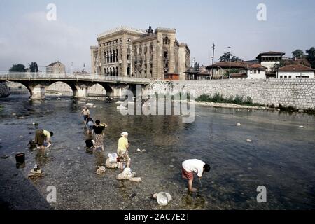 9 août 1993 pendant le siège de Sarajevo : les femmes laver la lessive dans la rivière Miljacka, avec l'holocauste de National Art Gallery et de la bibliothèque à l'arrière-plan. Aujourd'hui, la Bibliothèque est l'Hôtel de ville de Sarajevo. Banque D'Images