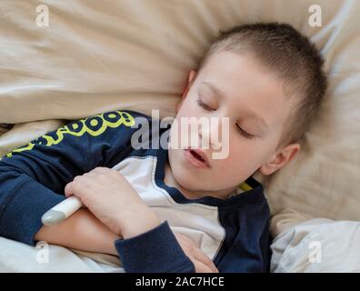 L'école maternelle garçon malade en pyjama couché sur un oreiller dans le lit, tenant en main un thermomètre numérique. Mauvais Garçon est la mesure de la température du corps et ne me sens pas bien Banque D'Images