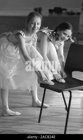 Années 1980, historiques, deux jeunes filles dans les costumes de danse à un ballet ou d'école de danse, avec l'aide d'un ballet de leurs pointes ou placé sur une chaise en plastique, England, UK. Les filles portent une pointe de la chaussure, un type de chaussures portées par des danseurs de ballet lorsque la formation ou l'exécution, qu'elles permettent aux danseurs de danser sur la pointe des pieds pendant de longues périodes de temps. Banque D'Images