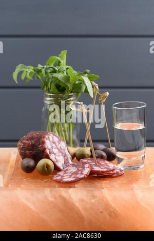 Shot de vodka avec amuse-gueules : salami fumé, roquette fraîche, verte et olives calamata situé sur hymalayan vertical bloc sel rose Banque D'Images