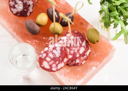 Salami, olives calamata et vert, roquette fraîche comme apéritif servi avec de la vodka tourné sur bloc de sel hymalayan rose sur fond blanc vue aérienne Banque D'Images