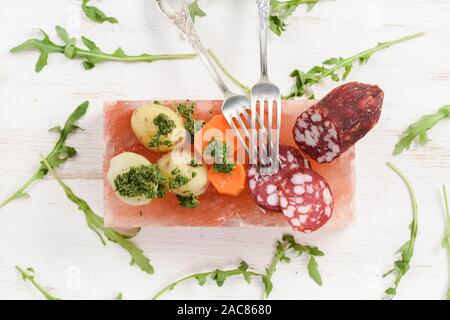 Salami, pommes de terre, carottes et de roquette fraîche avec sauce Chimichurri vert servi sur bloc de sel hymalayan rose sur fond blanc vue aérienne Banque D'Images