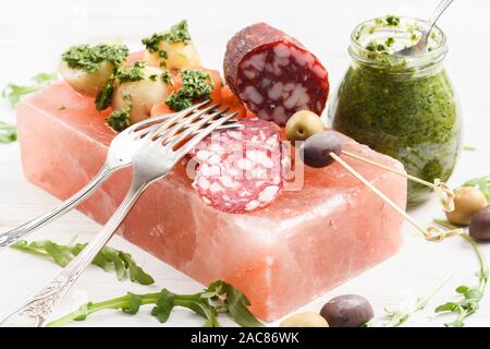 Apéritifs sur bloc de sel rose. Salami, pommes de terre, roquette, olives calamata et vert avec sauce Chimichurri vert vintage et fourchettes sur fond blanc Banque D'Images