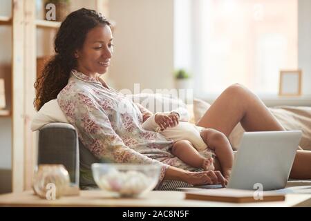 Vue de côté portrait de jeune mère afro-américaines à l'aide d'ordinateur portable de câliner bébé endormi dans la lumière du soleil, copy space Banque D'Images