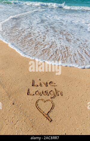 Les mots vivre, rire, et un coeur écrit dans le sable sur la plage avec une vague de lave Banque D'Images