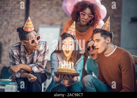 Faire un souhait. Dark-haired girl blowing anniversaire bougies et faire un vœu assis près de friends Banque D'Images