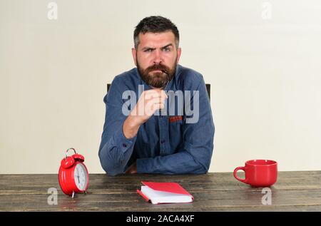 Homme à barbe est titulaire des verres, isolé sur fond blanc. Enseignant avec visage sérieux est assis à table en bois. Tasse, réveil rétro et vintage rouge livres sur table. Examen et étude de concept Banque D'Images